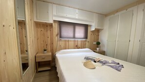 A bedroom with a bed, wooden walls, a small bedside table with a plant, a window with blinds, and a wardrobe. - Camping Arena Blanca (Benidorm)