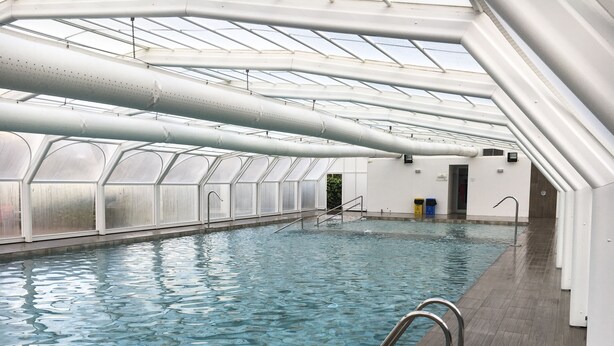 An indoor swimming pool with clear water, surrounded by a high ceiling with skylights and white structural beams. - Camping Arena Blanca (Benidorm)