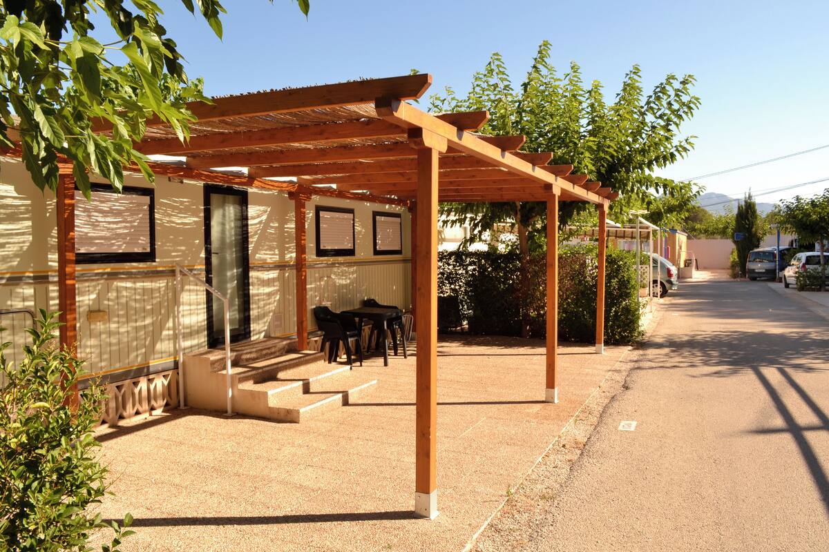 Ein überdachter Außenbereich mit Tisch und Stühlen, einer Pergola und einem Gebäude mit Tür und Fenstern.