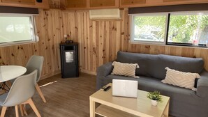 A cozy room with a grey sofa, a small table with a laptop, a dining area with chairs, and a wood-paneled wall with a window. - Camping Arena Blanca (Benidorm)