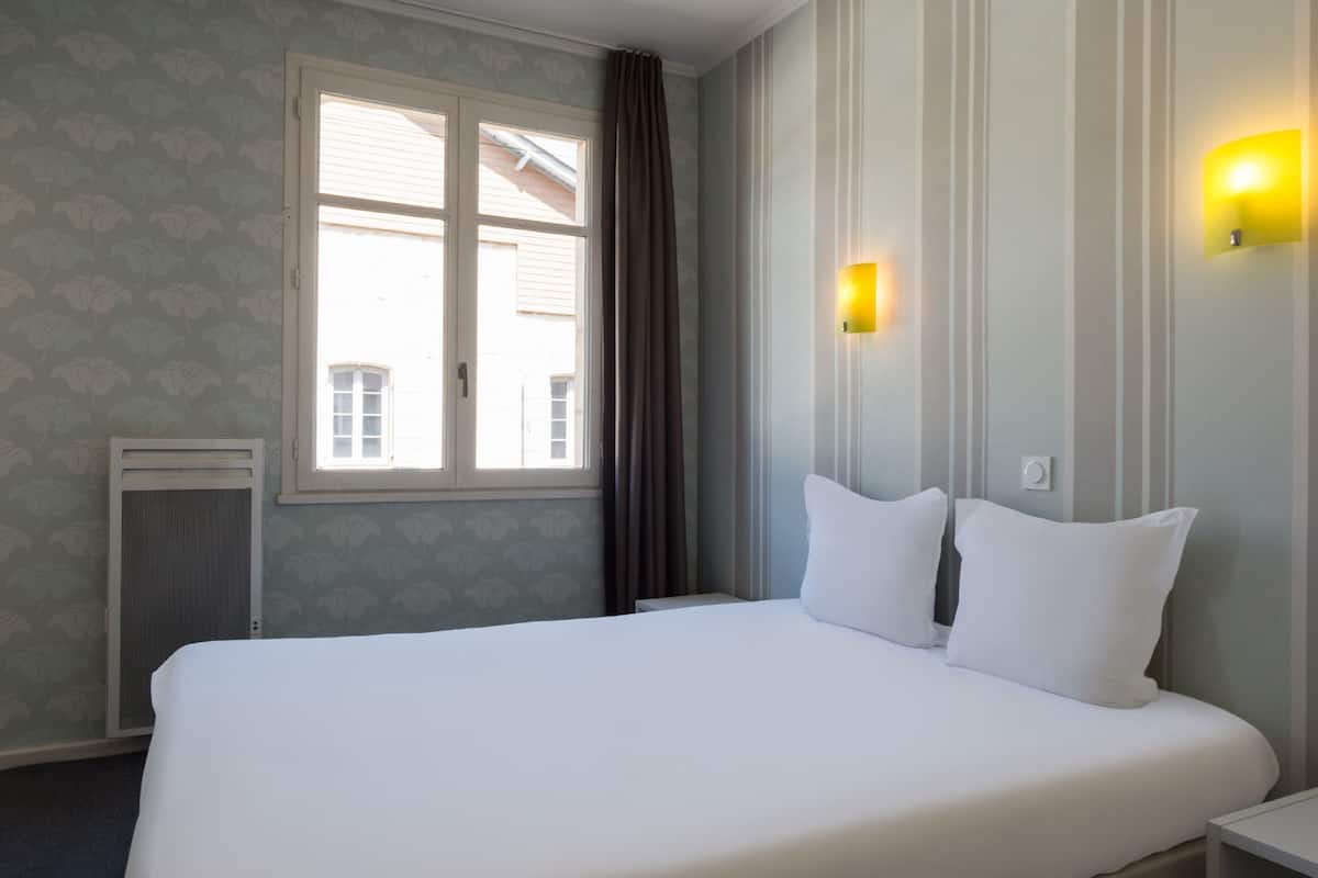 A hotel room with a large bed, a window with curtains, a wall-mounted light, and a radiator.
