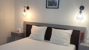 Una cama bien tendida con sábanas blancas y dos toallas enrolladas, un cuadro en la pared y dos lámparas de noche.