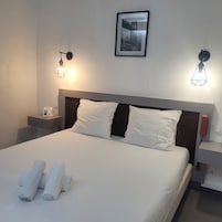 Una cama bien tendida con sábanas blancas y dos toallas enrolladas, un cuadro en la pared y dos lámparas de noche.