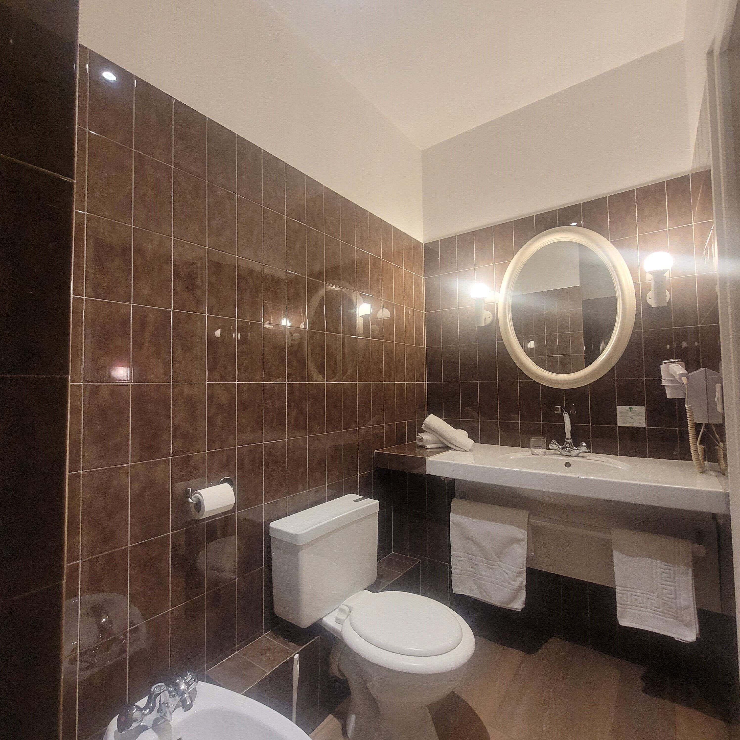 Une salle de bain moderne avec des carreaux de couleur marron foncé, des toilettes blanches, un lavabo avec un miroir et des serviettes.