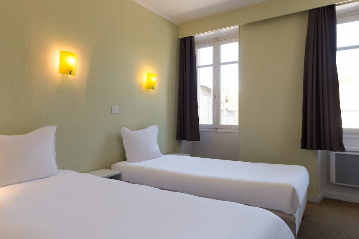 A hotel room with two single beds, a window with curtains, and two wall-mounted light fixtures.