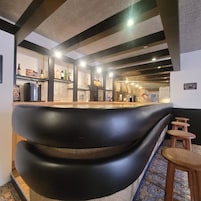 Una zona de bar con una barra negra curva, estantes con botellas, vasos y un cartel en la pared.
