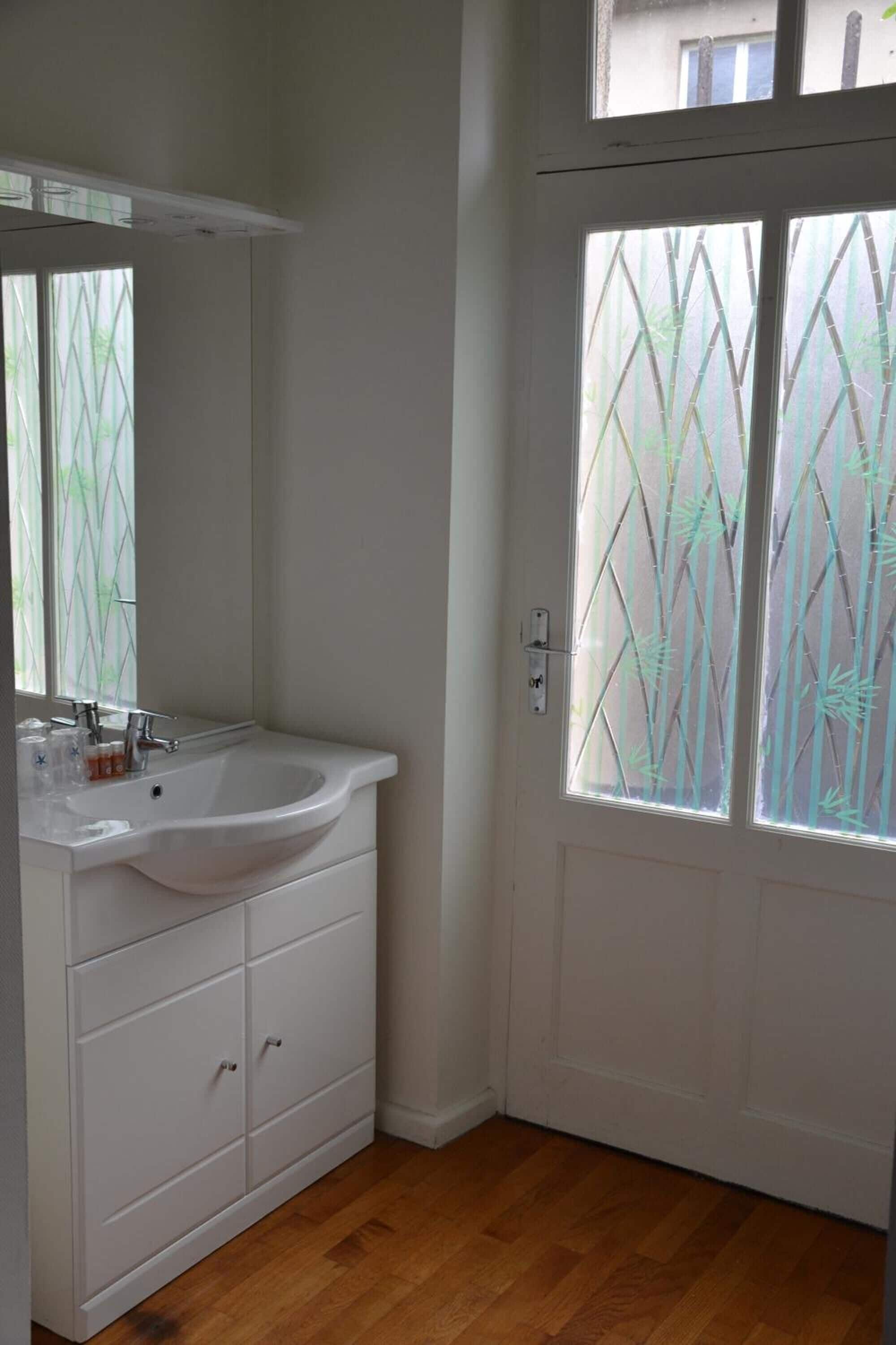 A bathroom with a white sink, mirror, and cabinet, a frosted glass door, and wooden flooring.