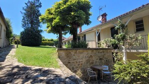 Un camino de piedra que lleva a un muro de piedra con una mesa y sillas, rodeado de follaje y un árbol grande.
