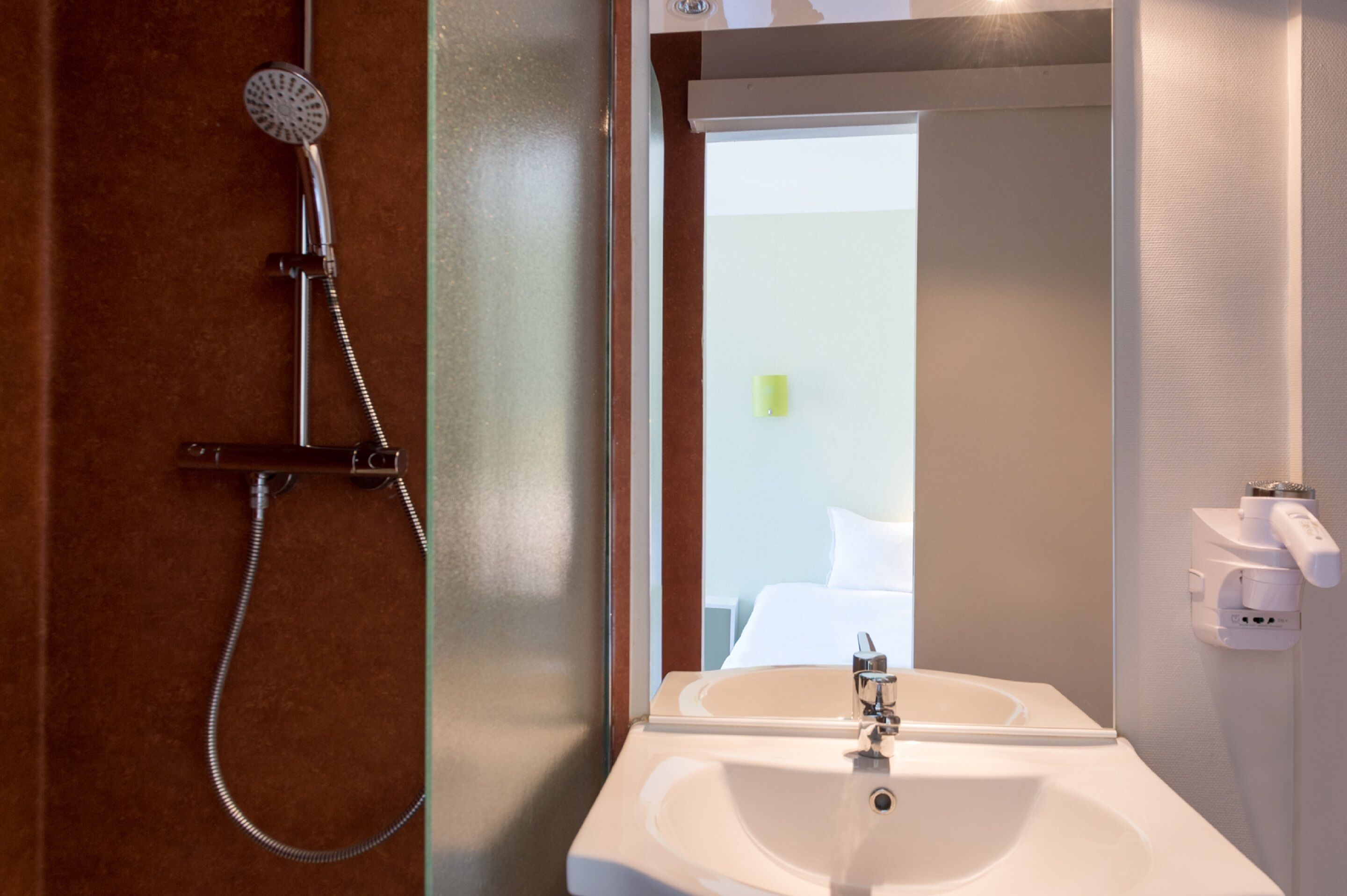 A bathroom with a shower, sink, and a mirror reflecting a bed and a wall-mounted light fixture.