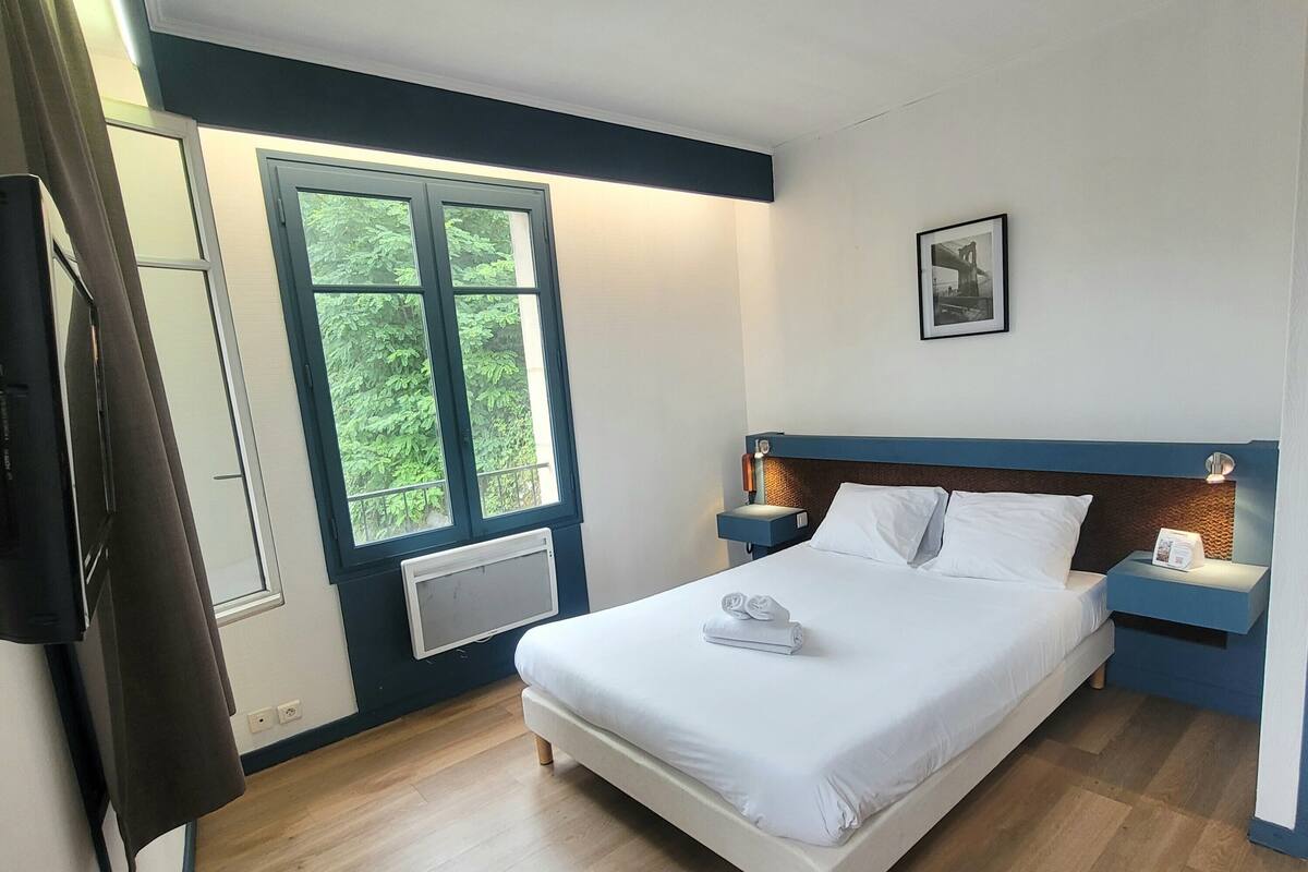 A hotel room with a bed, a nightstand, a window with a view of greenery, and a wall-mounted picture.