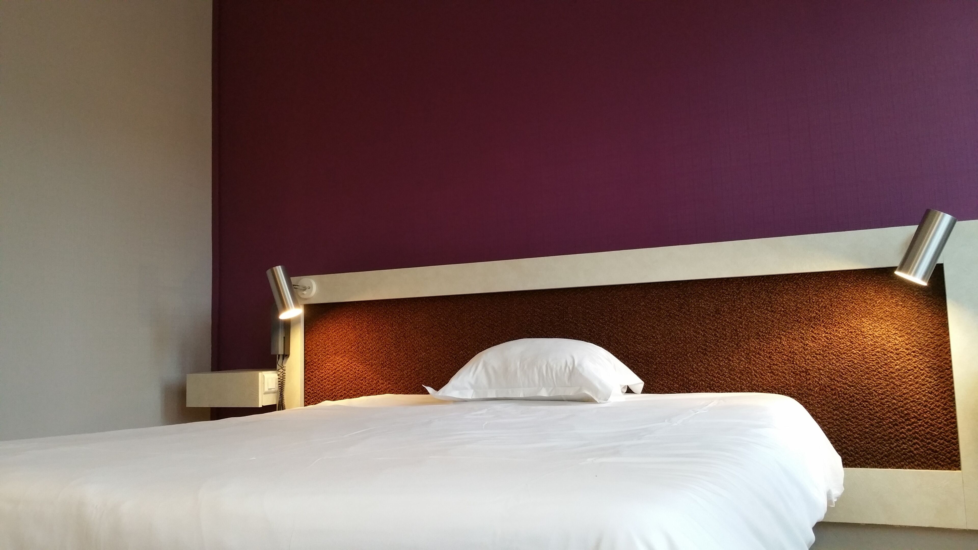 Un lit avec une literie blanche, une tête de lit ornée d’un panneau marron texturé et un mur violet.