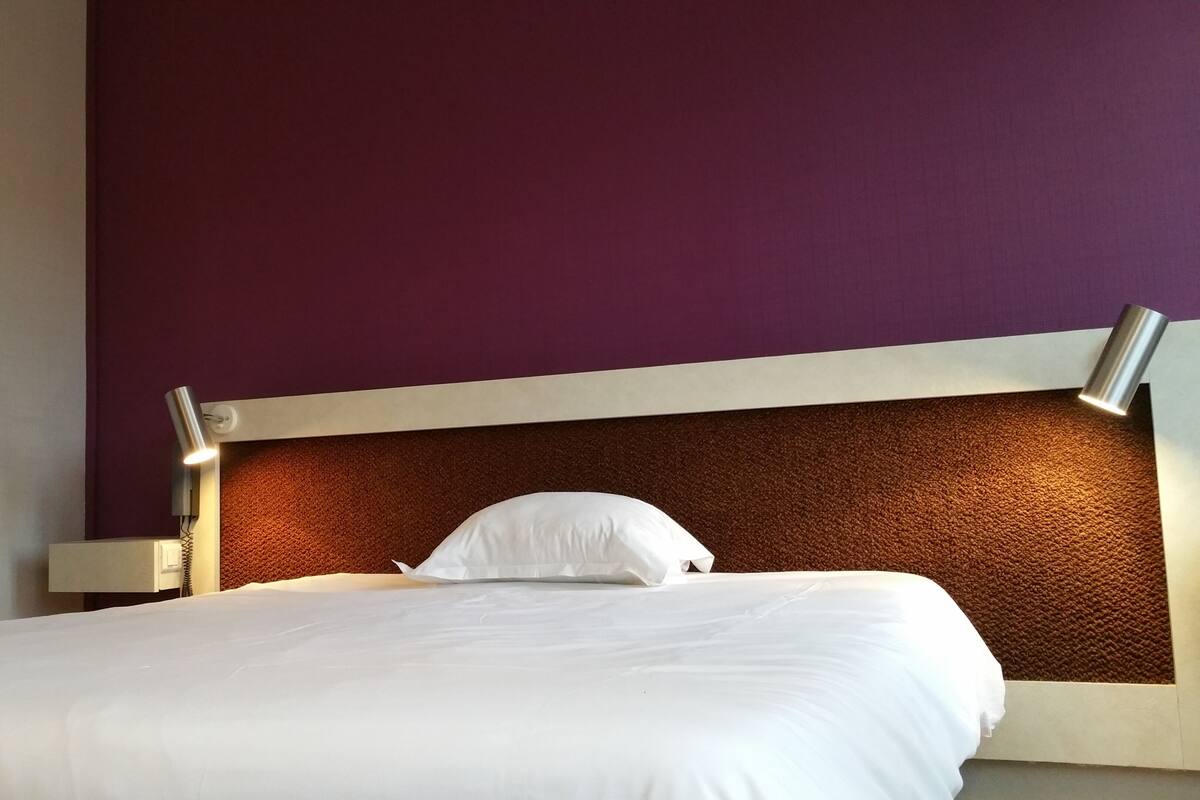 A bed with white bedding, a headboard with a textured brown panel, and a purple wall.