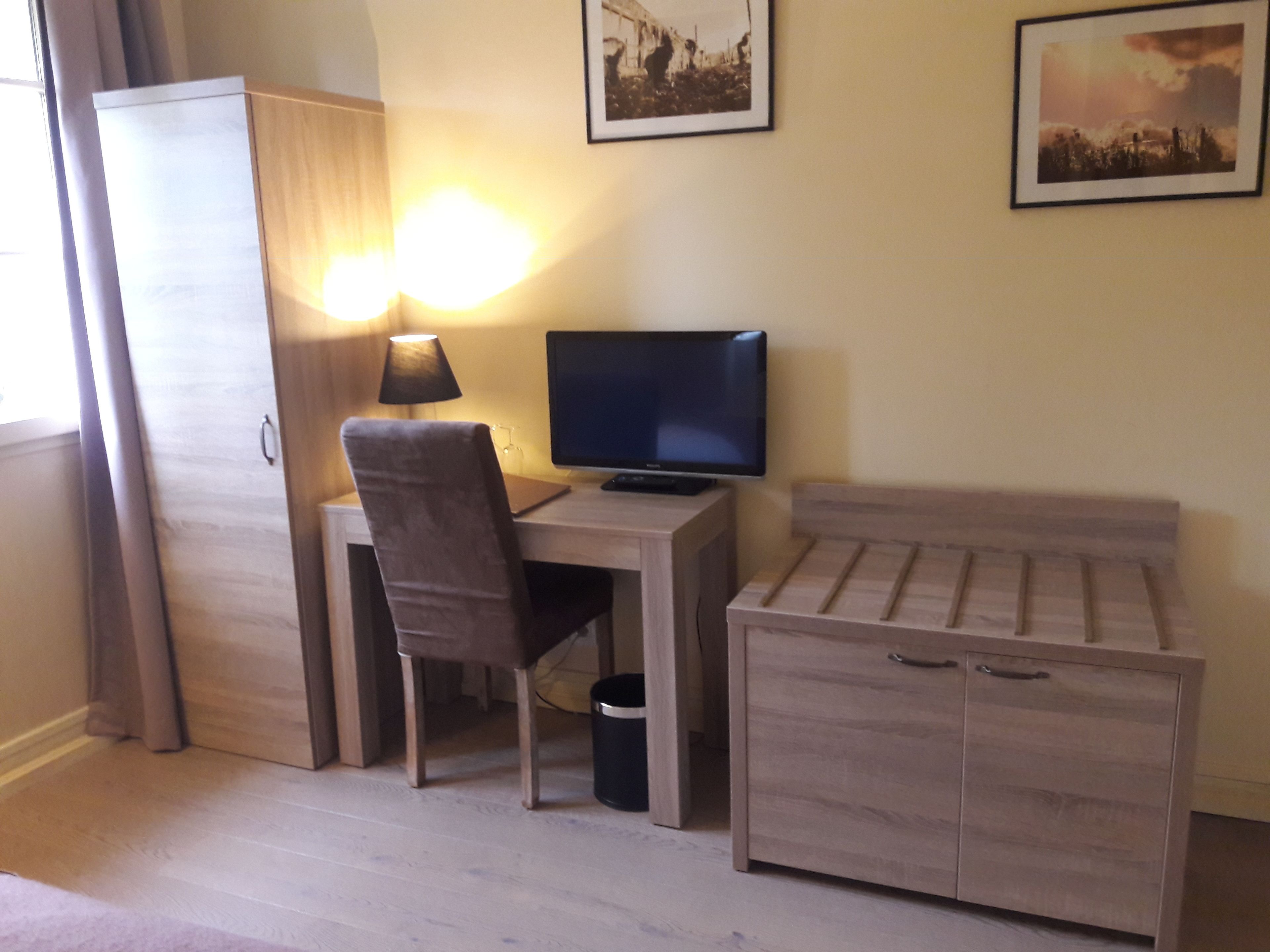 A small, well-lit room with a desk, chair, television, and a bed.