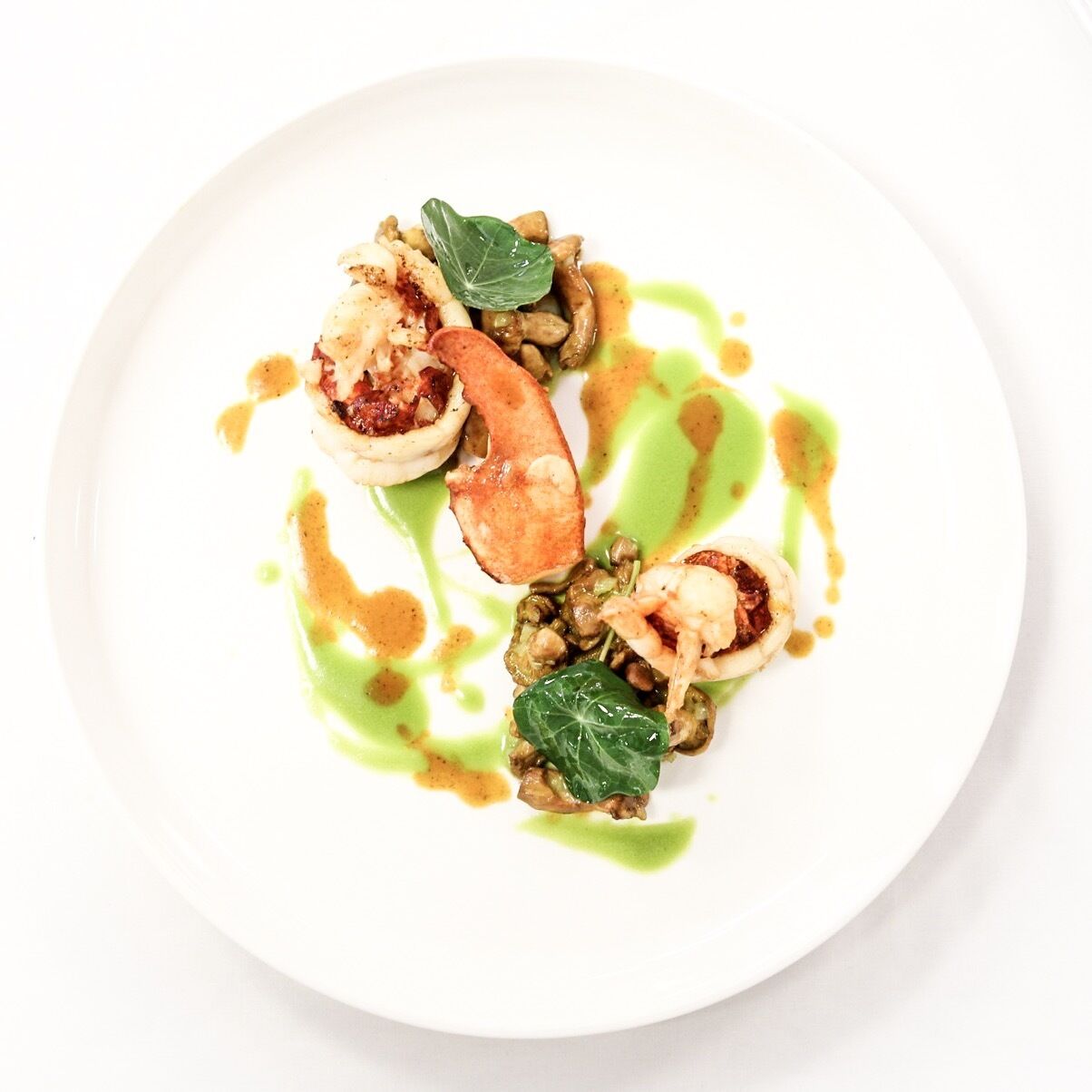 Une assiette blanche contenant un plat de fruits de mer, notamment des moules et un morceau de poisson, agrémenté d’une sauce verte et d’une feuille.