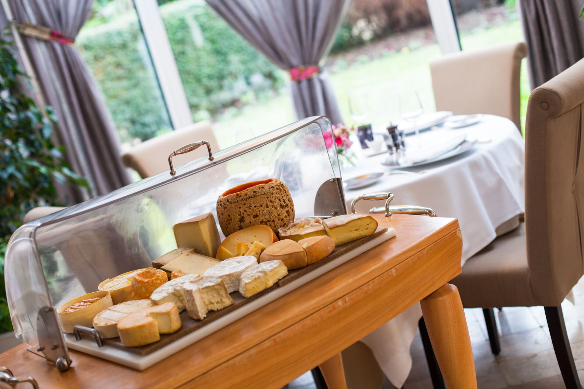 A cheese board with various types of cheese and bread on a wooden table in a room with a view of greenery outside.
