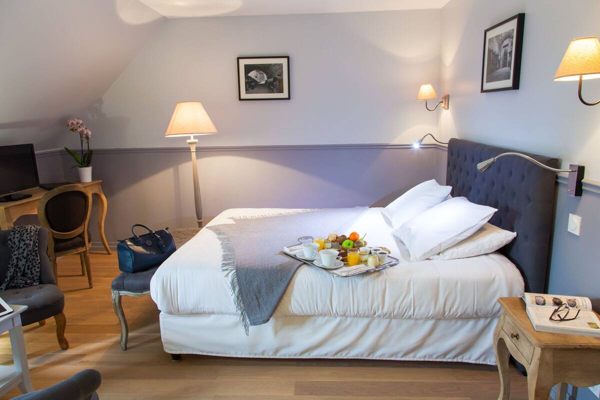 Habitación de hotel ordenada con cama, mesita de noche, lámpara, silla y un pequeño escritorio con una computadora portátil.