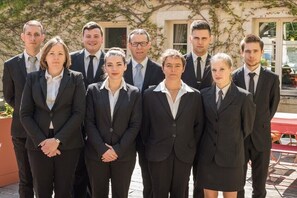A group of nine people in business attire standing in front of a building with a red table and chairs in the background.