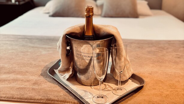 A bed with a champagne bottle and glasses on a tray, a nightstand with a lamp, and a headboard with pillows. - Hôtel Saint Martin (Orléans)
