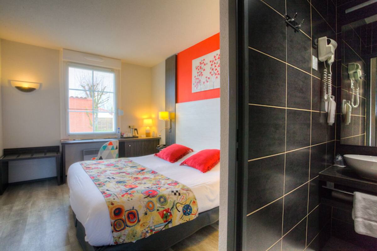A hotel room with a bed, a desk, a window, and a bathroom with a sink and a hairdryer.