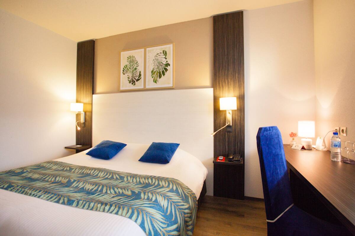 A hotel room with a large bed, two blue pillows, a desk with a chair, and two wall-mounted lamps.