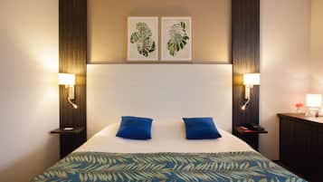 Ein Hotelzimmer mit einem großen Bett, zwei blauen Kissen, einer gemusterten Bettdecke, zwei Wandleuchten und zwei gerahmten Blätter-Kunstwerken.
