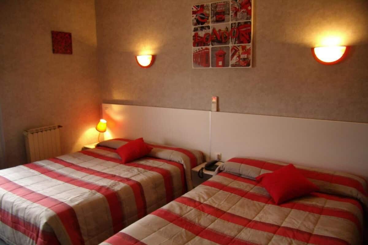 A room with two beds, each with a red and beige checkered bedspread, a small bedside table, and a wall-mounted light fixture.