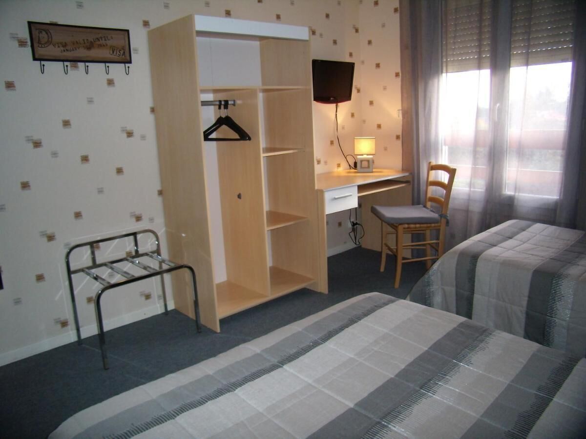 A hotel room with a bed, a desk with a chair, a TV mounted on the wall, a wardrobe, and a window with curtains.