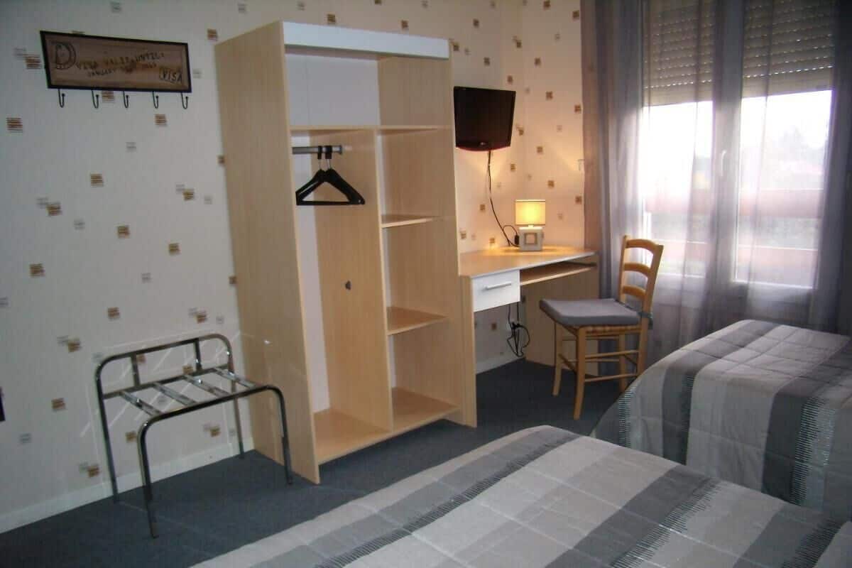 A hotel room with a bed, a desk with a chair, a TV mounted on the wall, a wardrobe, and a window with curtains.