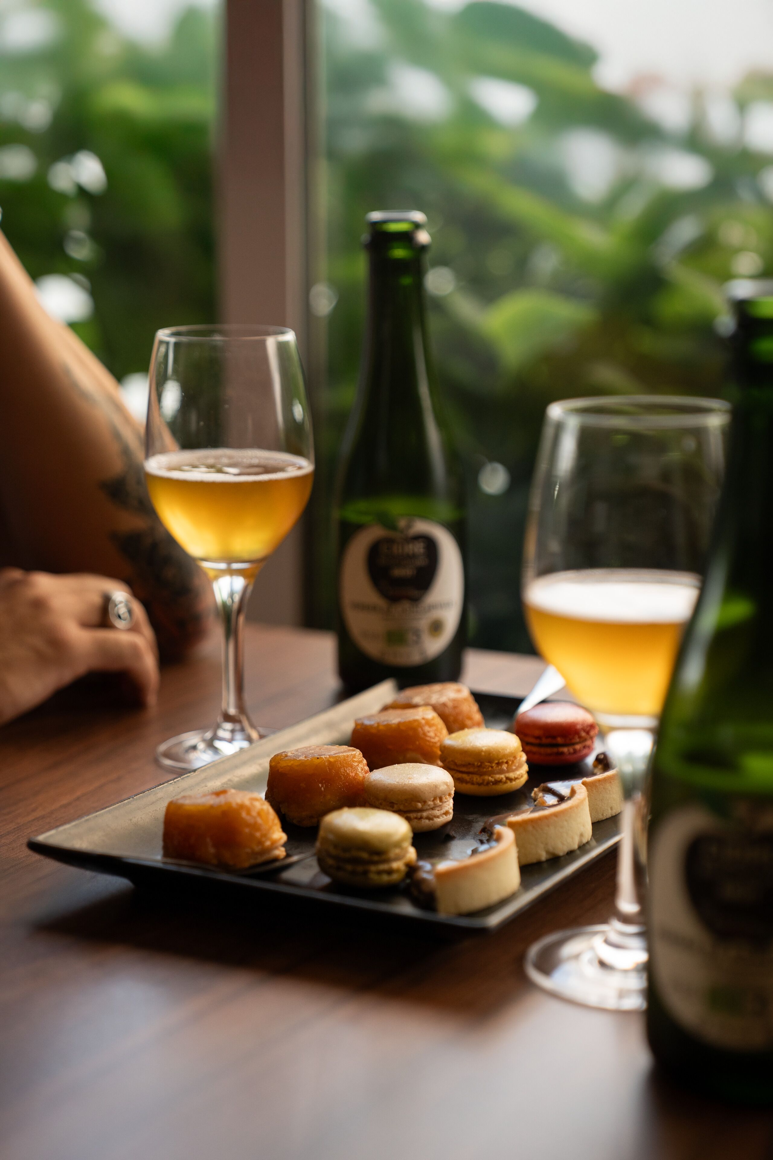Una bandeja con pasteles variados y dos vasos de cerveza sobre una mesa, con una botella de vino al fondo.