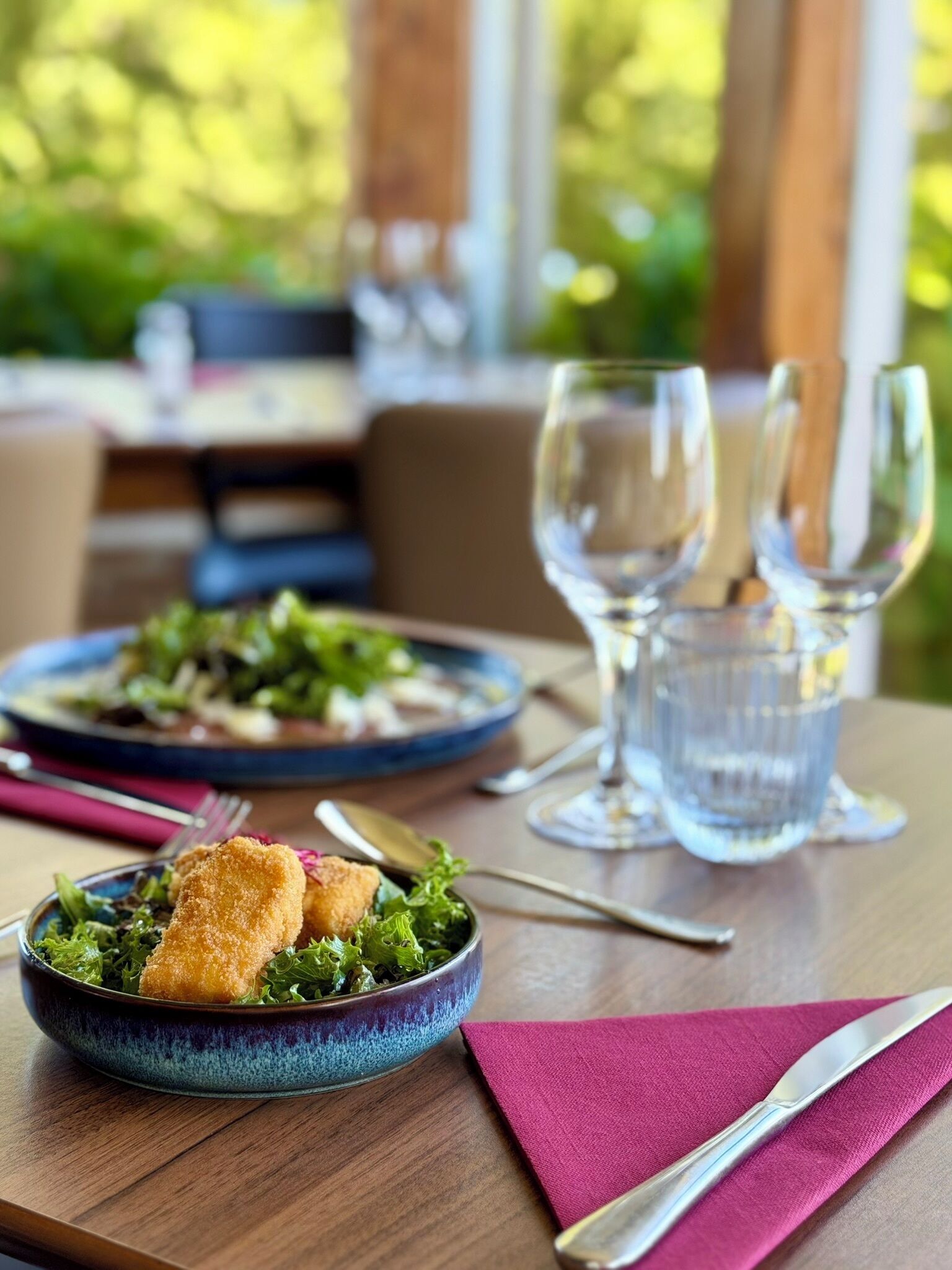 A dining table with a plate of food, a bowl of salad, and two wine glasses.