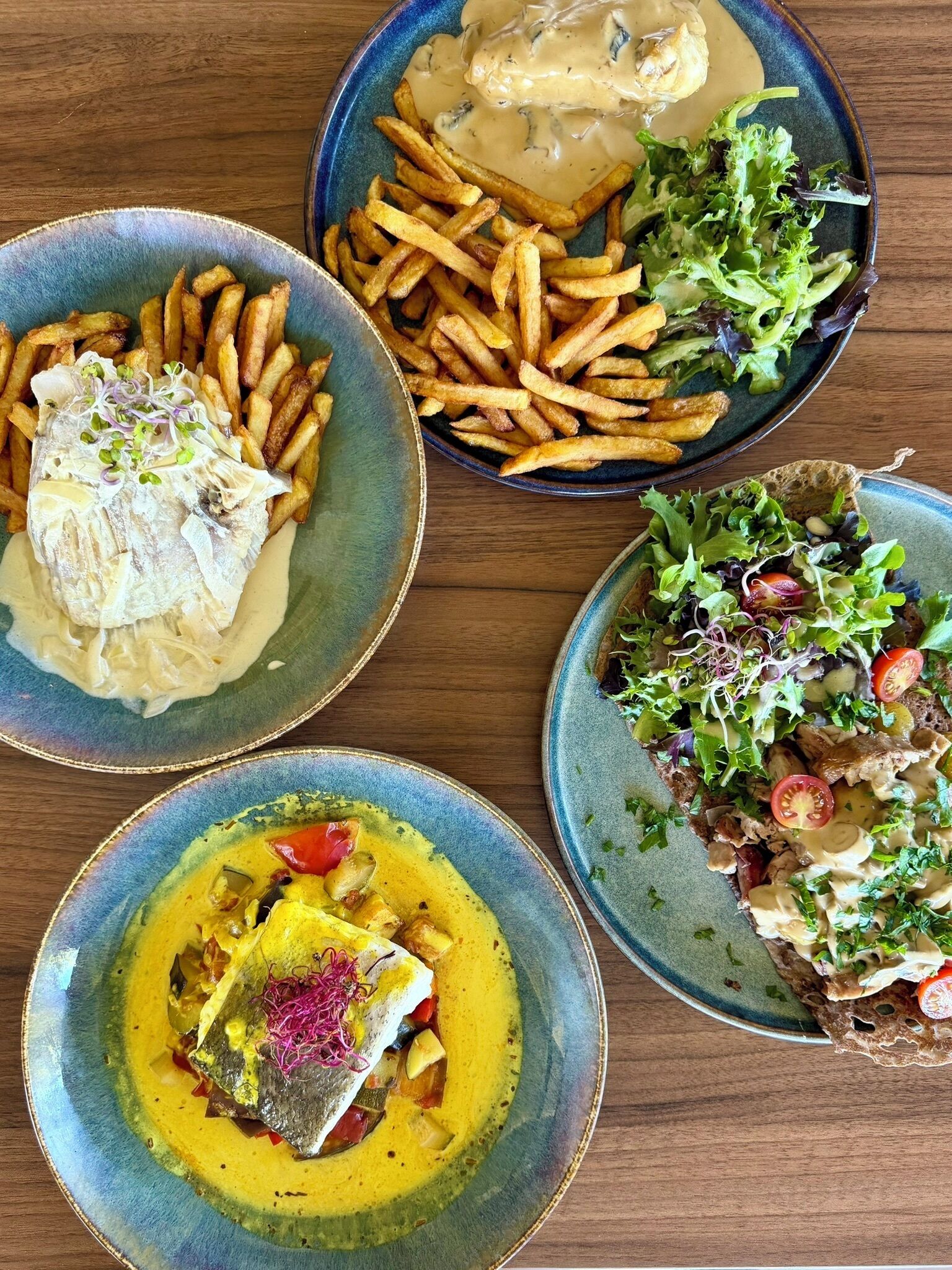 A table with four dishes: fish with sauce, fries with sauce, a salad, and a fish dish with vegetables.