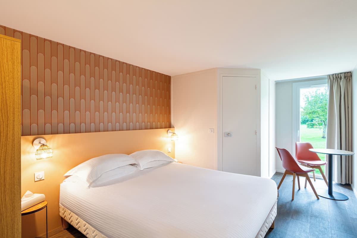 A hotel room with a large bed, two bedside lamps, a small table with chairs, and a view of the outdoors through a window.