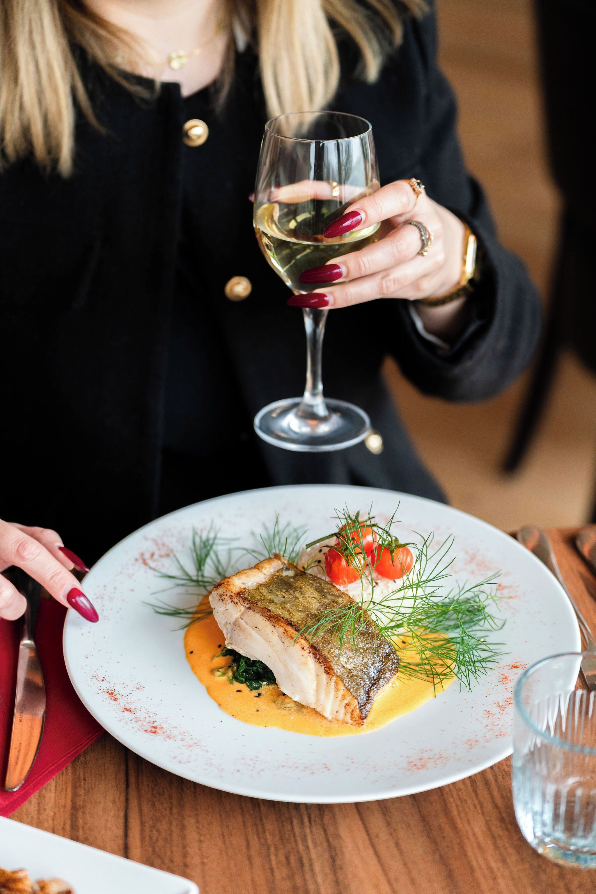 A person holding a glass of white wine, seated at a table with a plate of food.