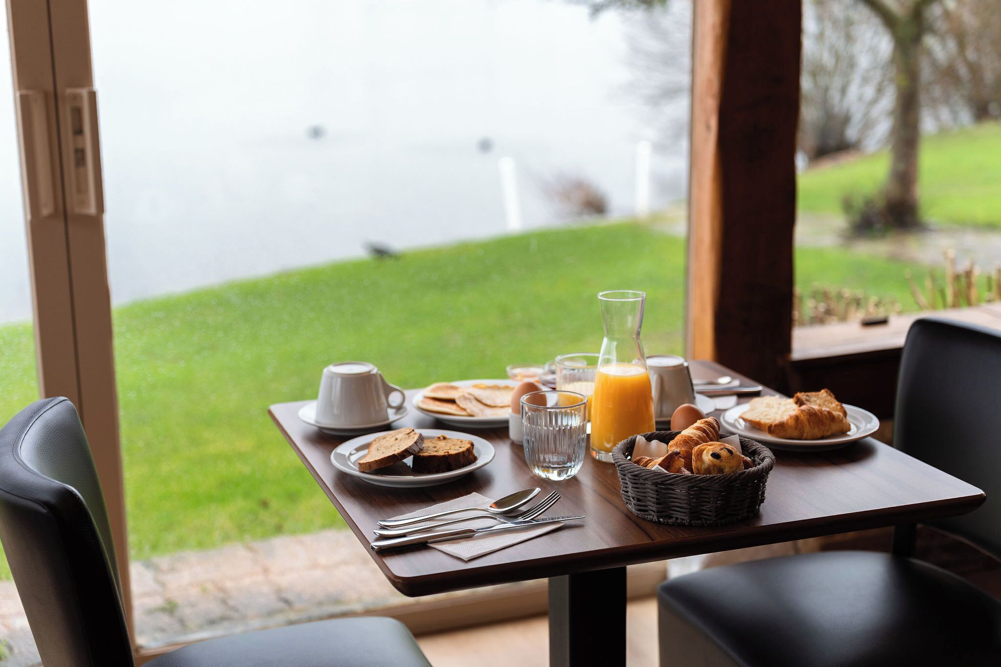 Una mesa de desayuno con platos de comida, un vaso de jugo y vistas a una zona verde y al agua.