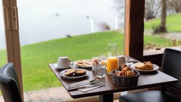 Una mesa de desayuno con platos de comida, un vaso de jugo y vistas a una zona verde y al agua.
