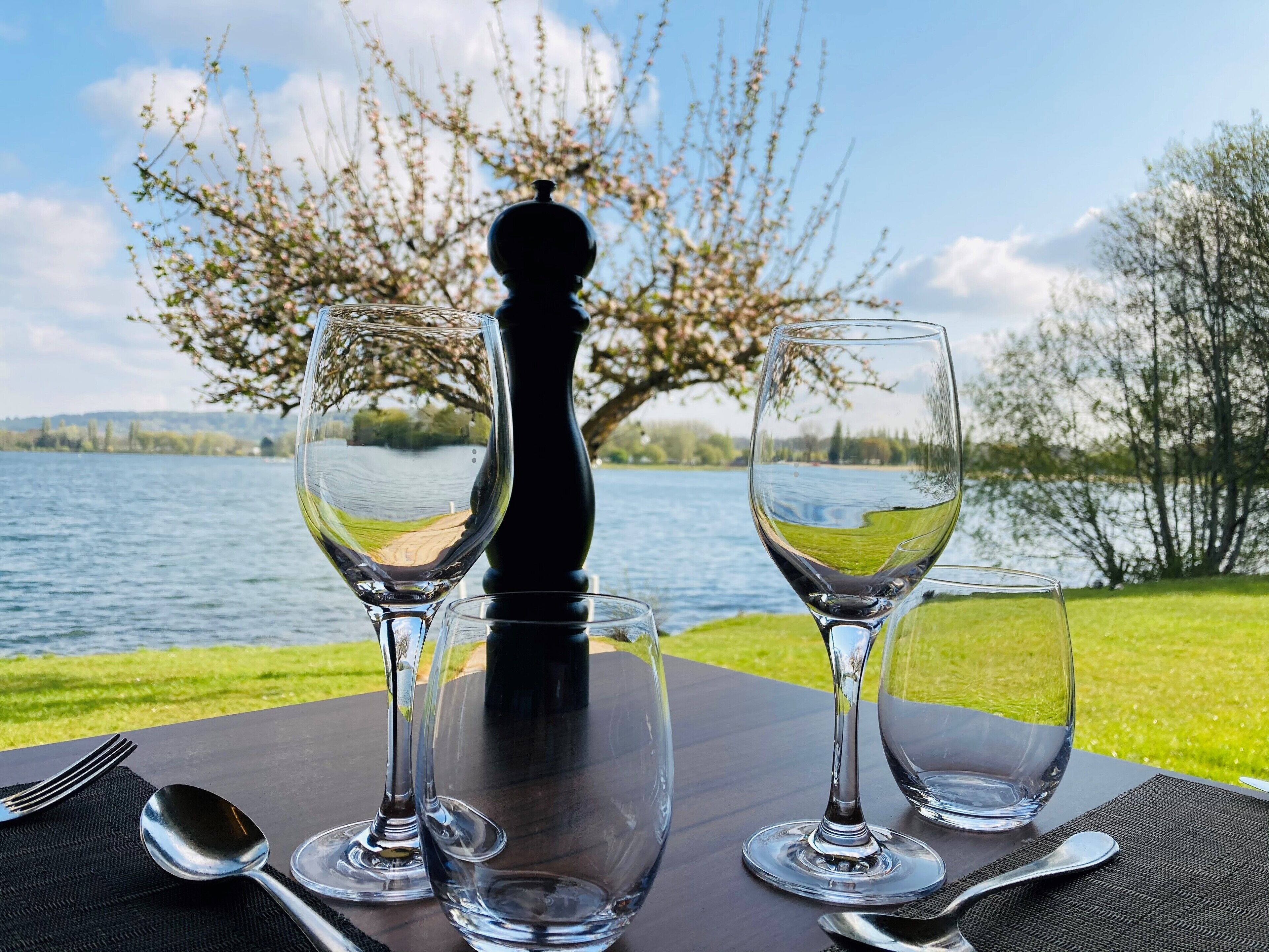 A table with three wine glasses and a spoon, set against a backdrop of a lake and trees.