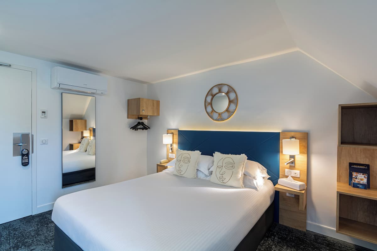 A hotel room with a large bed, a mirror, a headboard, bedside lamps, a wall-mounted air conditioner, and a wooden shelf.