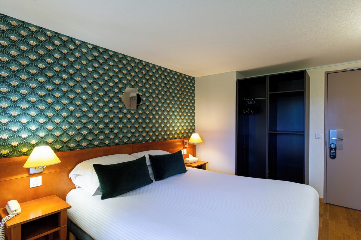 A hotel room with a bed, two bedside lamps, a nightstand, a telephone, a wall with a geometric pattern, and a closet.