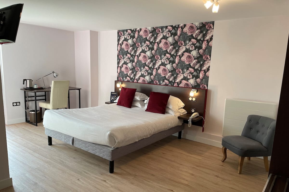 A hotel room with a bed, a desk, a chair, and a wall with a floral wallpaper.