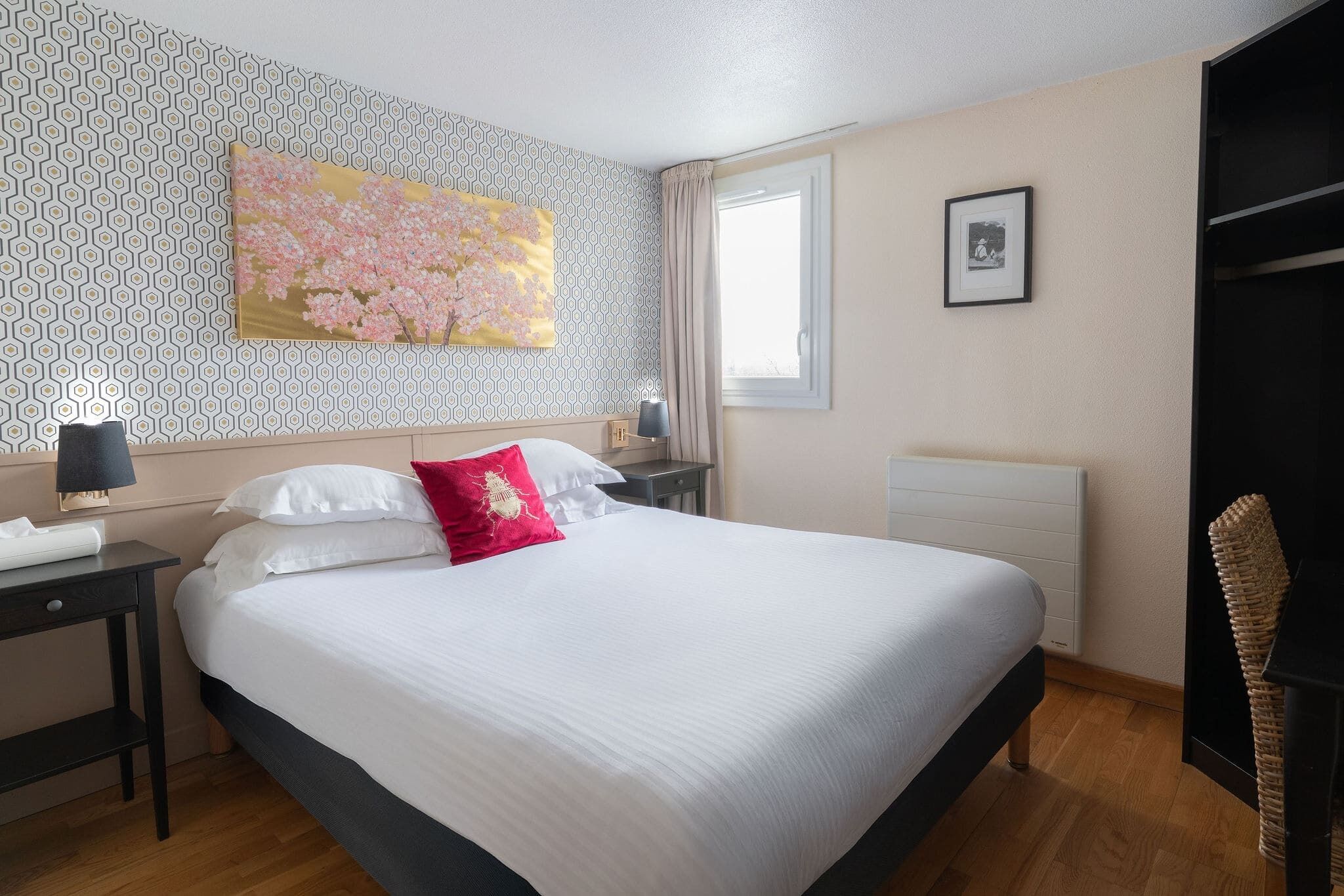 Une chambre à coucher bien rangée, avec un grand lit, une table de chevet avec une lampe, un tableau accroché au mur et une fenêtre avec des rideaux.
