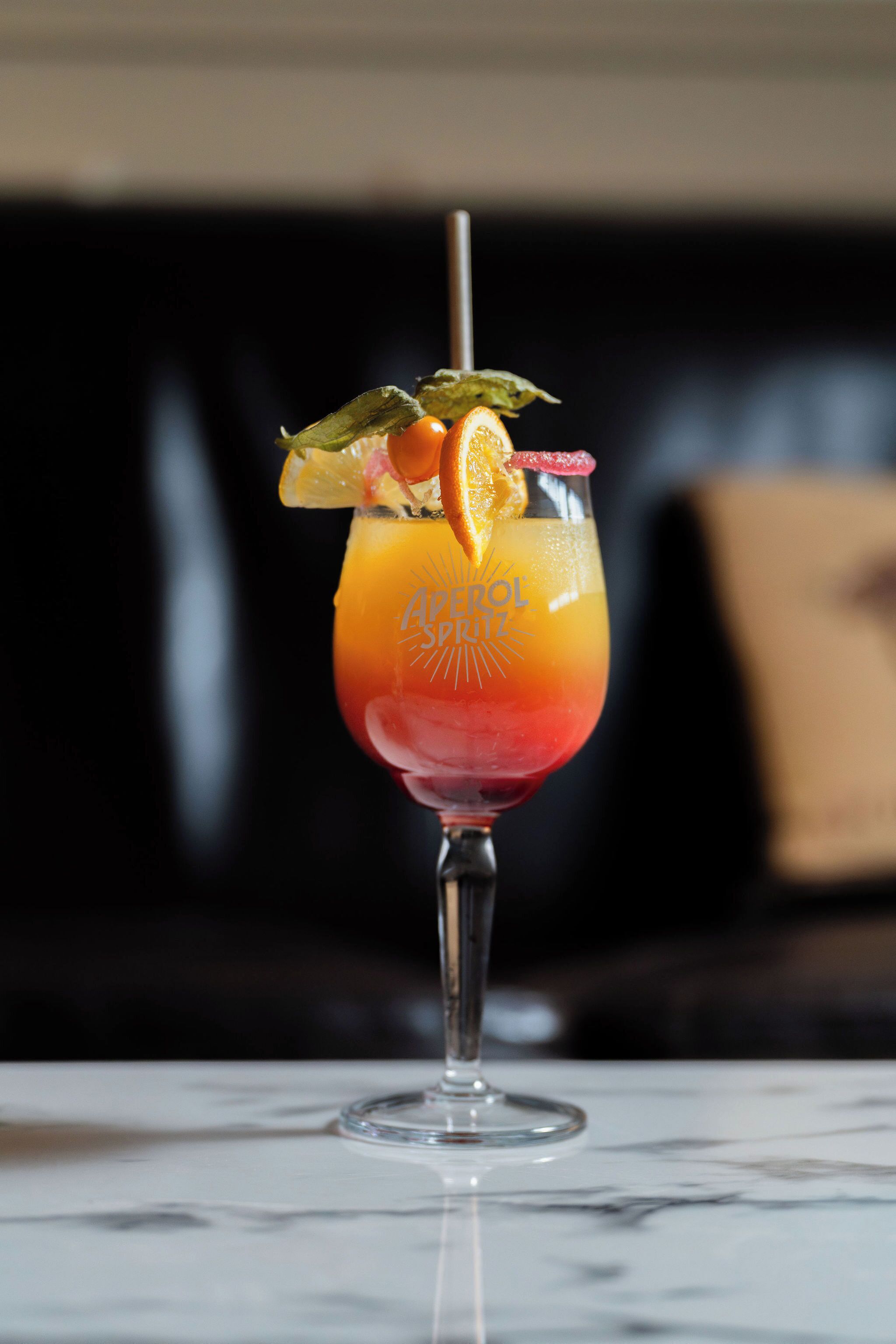 A cocktail in a stemmed glass with a straw, garnished with an orange slice and a green leaf, on a marble surface.