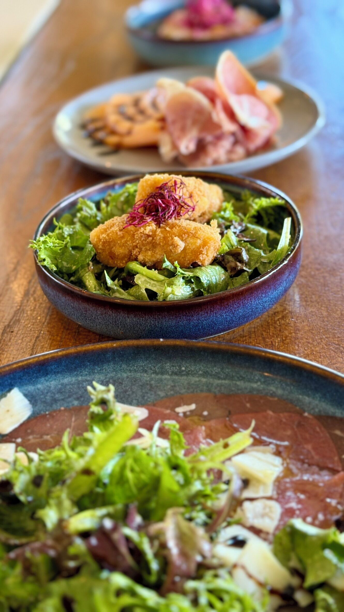 Een tafel met diverse gerechten, waaronder een salade met een goudbruin ingrediënt erop, een bord met plakjes vlees, en nog een bord met een andere salade.