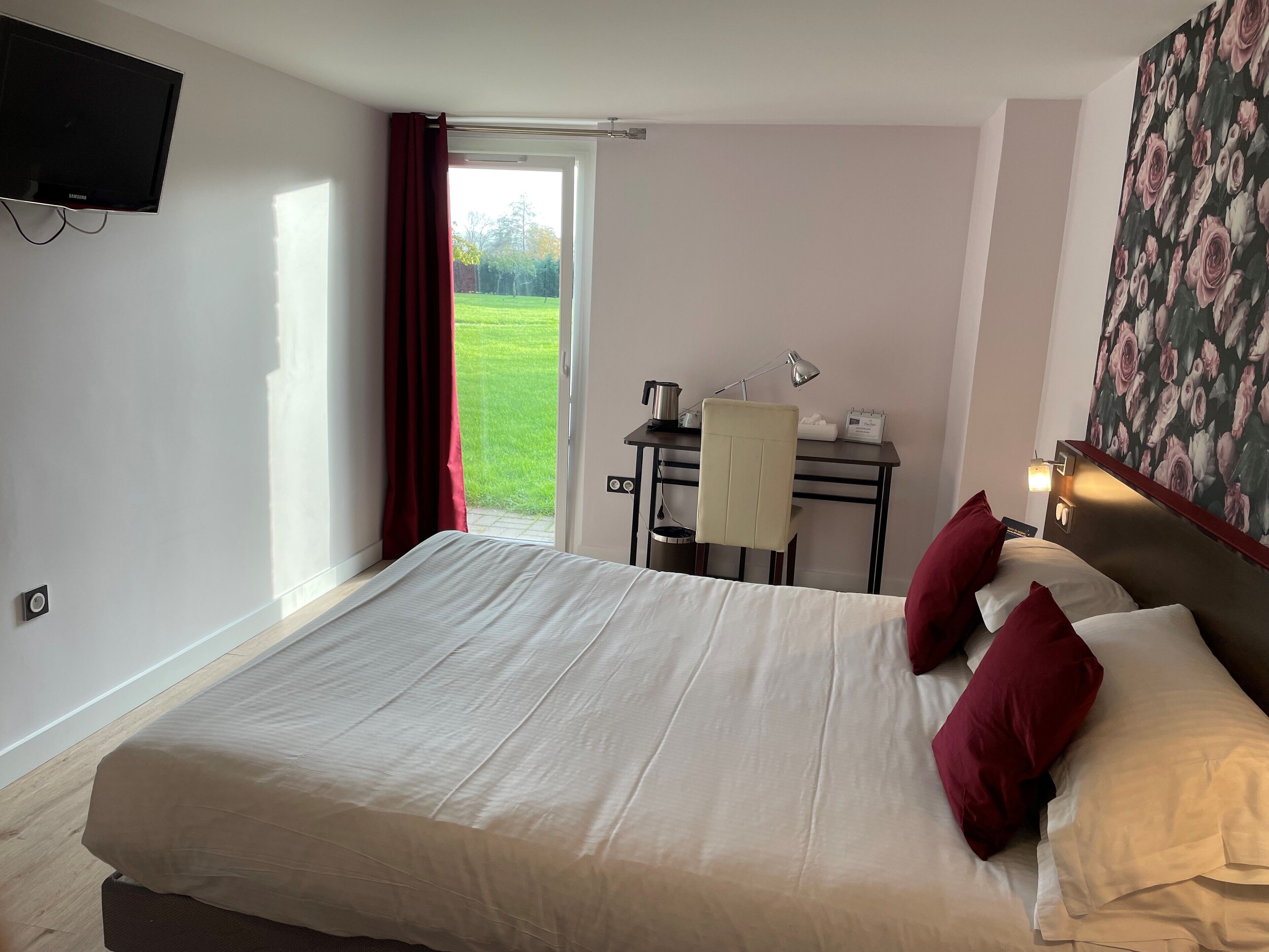 Une chambre d’hôtel avec un grand lit, un bureau avec une chaise, une télévision et une fenêtre donnant sur une pelouse verdoyante.