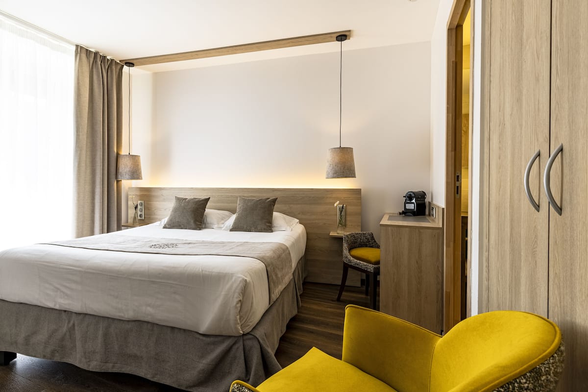 A modern hotel room with a large bed, a yellow armchair, a wooden headboard, and a small desk with a lamp.
