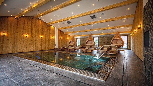 Una amplia piscina interior con paredes y techo de madera, rodeada de sillones y sillas colgantes.