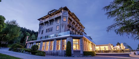 A multi-story hotel with a balcony on the upper floor, a gravel driveway, and outdoor seating area with umbrellas.