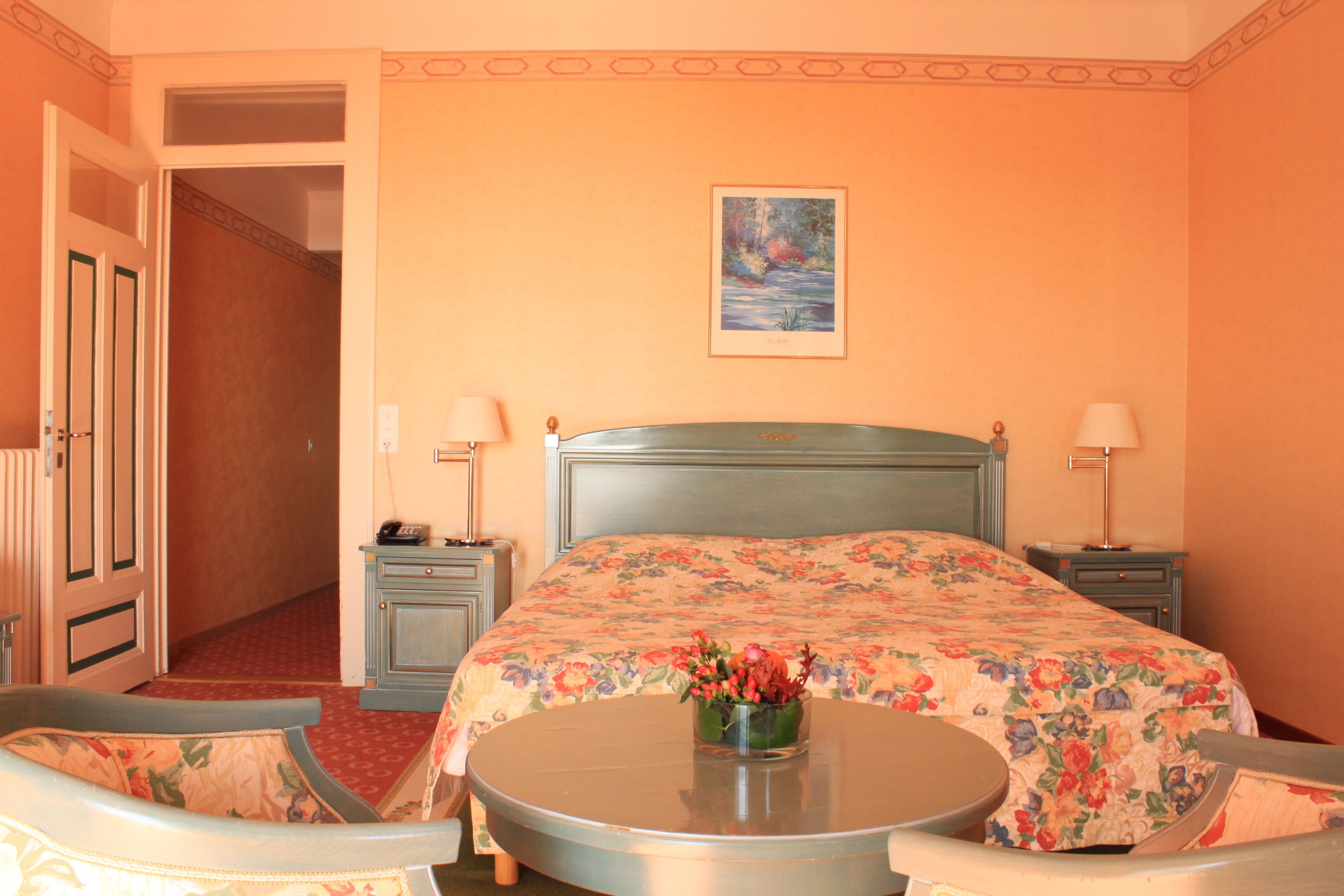 Habitación de hotel con una cama, dos mesitas de noche, una silla, una mesa redonda y un cuadro en la pared.