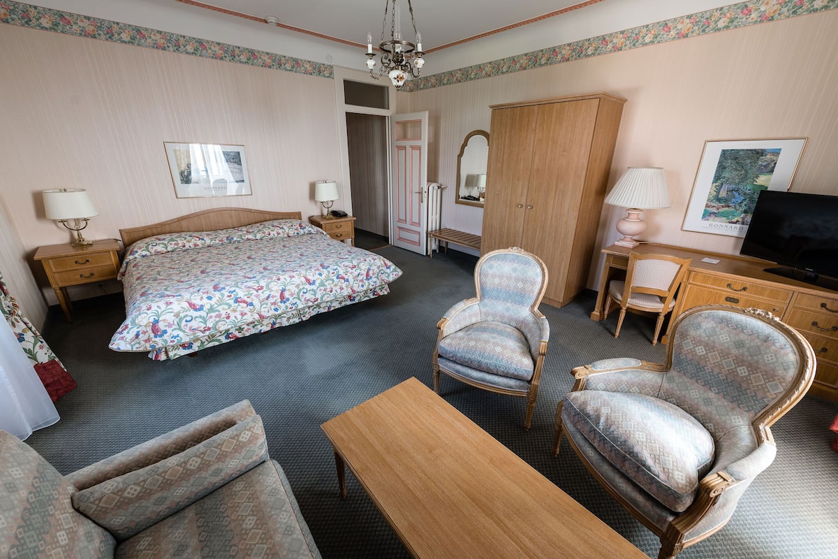 A hotel room with a bed, two armchairs, a wooden table, a desk with a chair, a television, and a wardrobe.