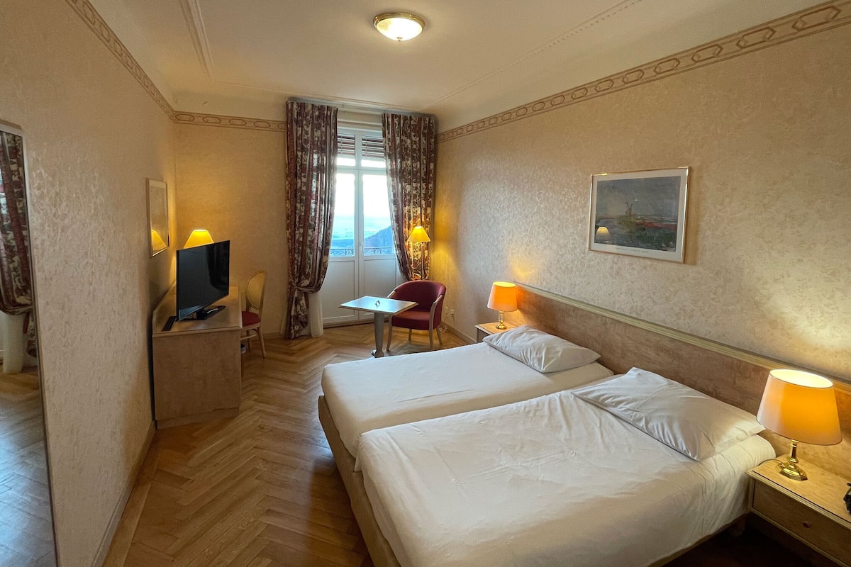 A hotel room with two beds, a desk, a chair, a TV, and a window with curtains.