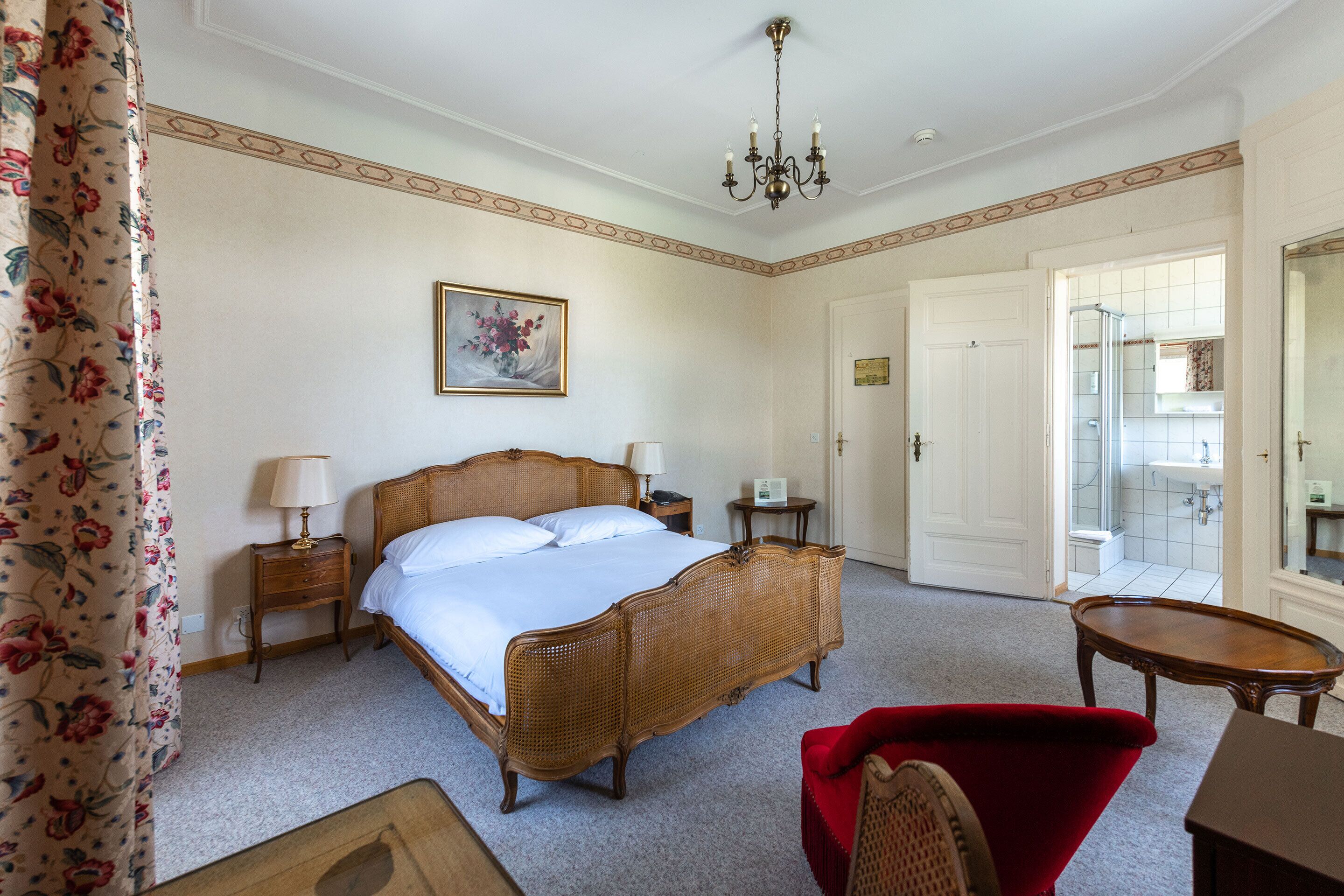 Une chambre à coucher avec un grand lit, deux tables de chevet, un fauteuil rouge et une salle de bain visible à travers une porte ouverte.